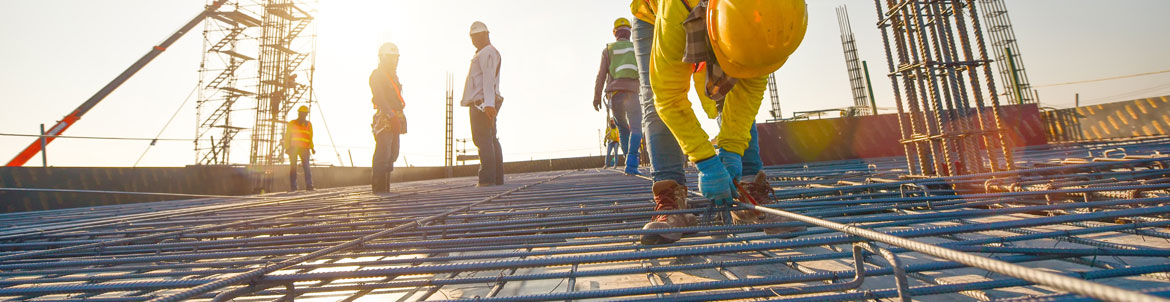 A construction worker is working on building structure in the foreground with five other construction workers standing in the background