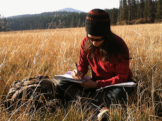 Woman sitting in brown grasses, writing on clipboard