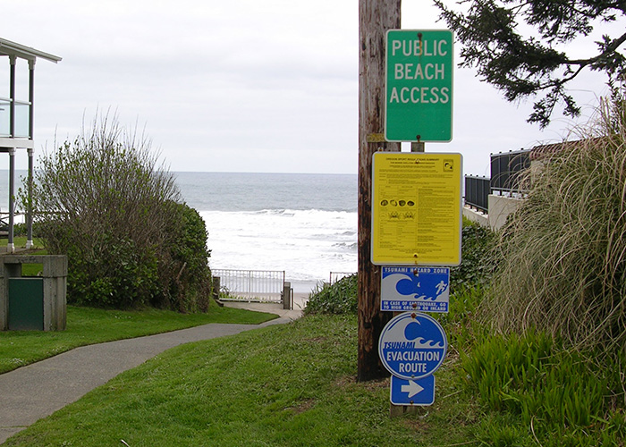 A sign by a beach path includes tsunami warnings and points towards an evacuation route