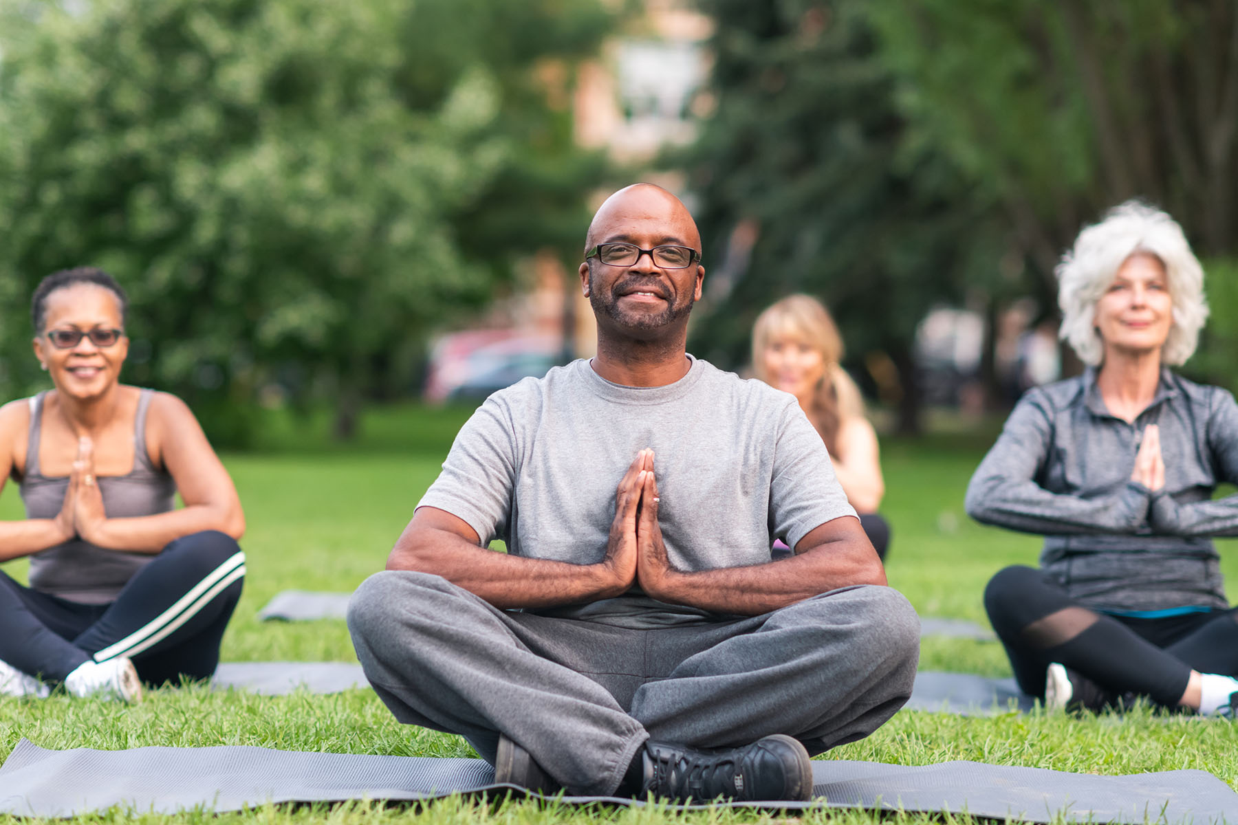 Multi-ethnic group of seniors meditating outdoors A multi-ethnic group of seniors is attending a yoga class outdoors. The group is sitting on yoga mats. They are meditating. The individual in focus is a black man. He is sitting at the front of the group. He is smiling directly at the camera. community exercise class stock pictures, royalty-free photos & images