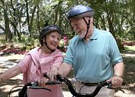 Two seniors, a man and a woman, wearing helmets and sitting on bikes in a park setting