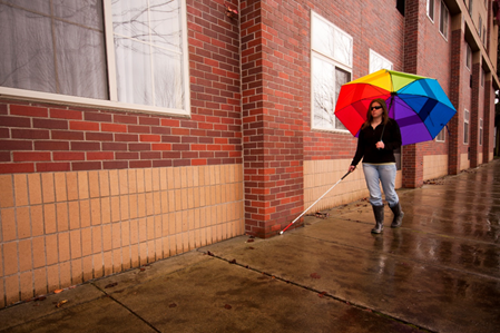 Woman with a white cane walks outdoors on a sidewalk.