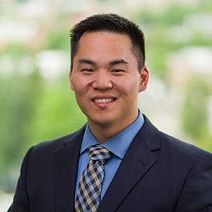 Eric, has brown short hair and is wearing a black suite jacket with a blue dress shirt and a checkered yellow and blue tie.