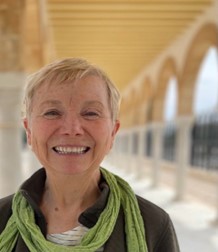 Jody, short blonde hair. Wearing a brown jacket with a white shirt and a green scarf.