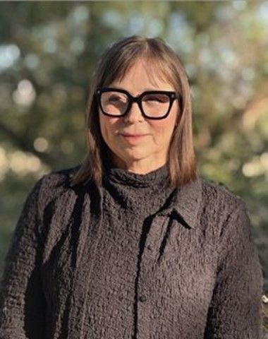 Sandra, shoulder length light brown hair, with thick black rimmed glasses. Wearing a brown textured jacket and a matching shirt.