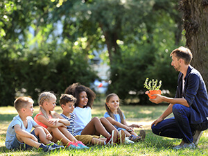 Teacher holding pot with plant in front of five children sitting in the grass