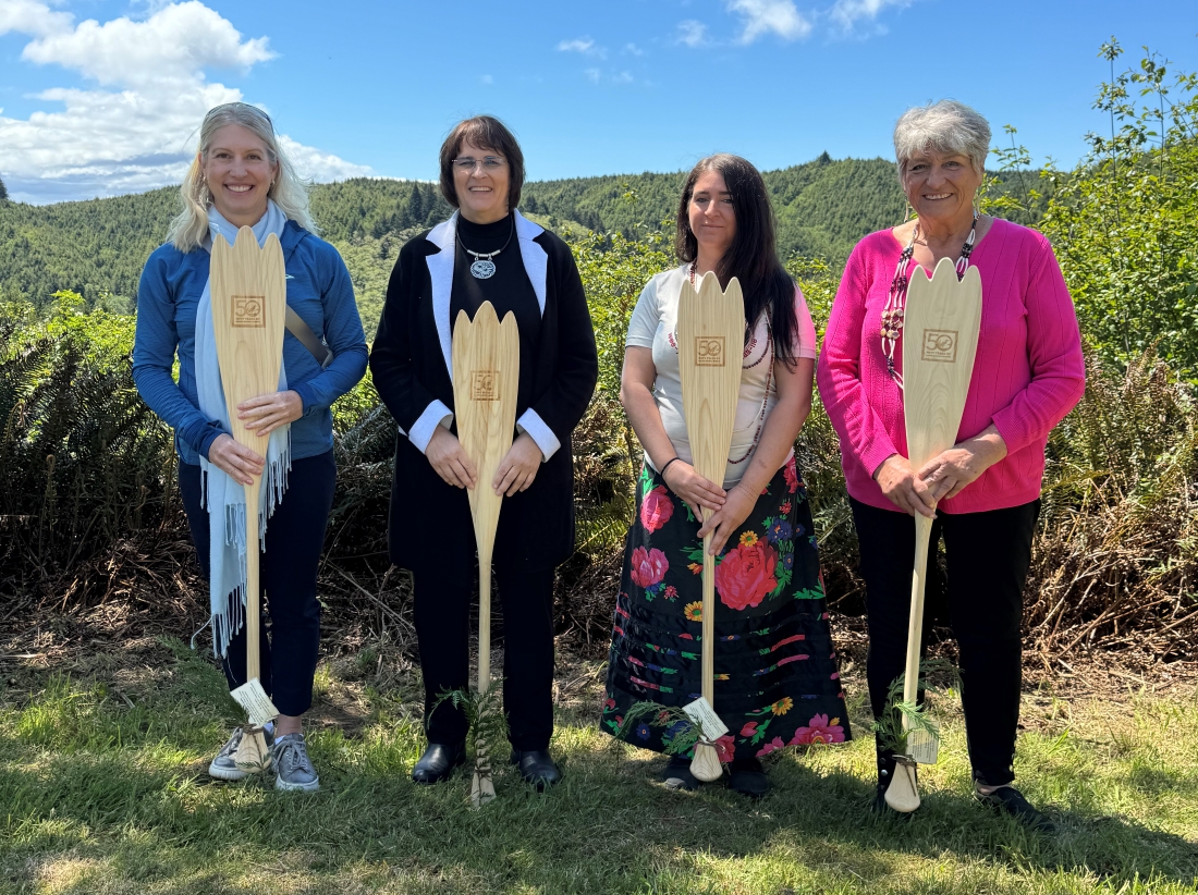 Four women smiling holding ceremonial paddles with a blue sky behind them