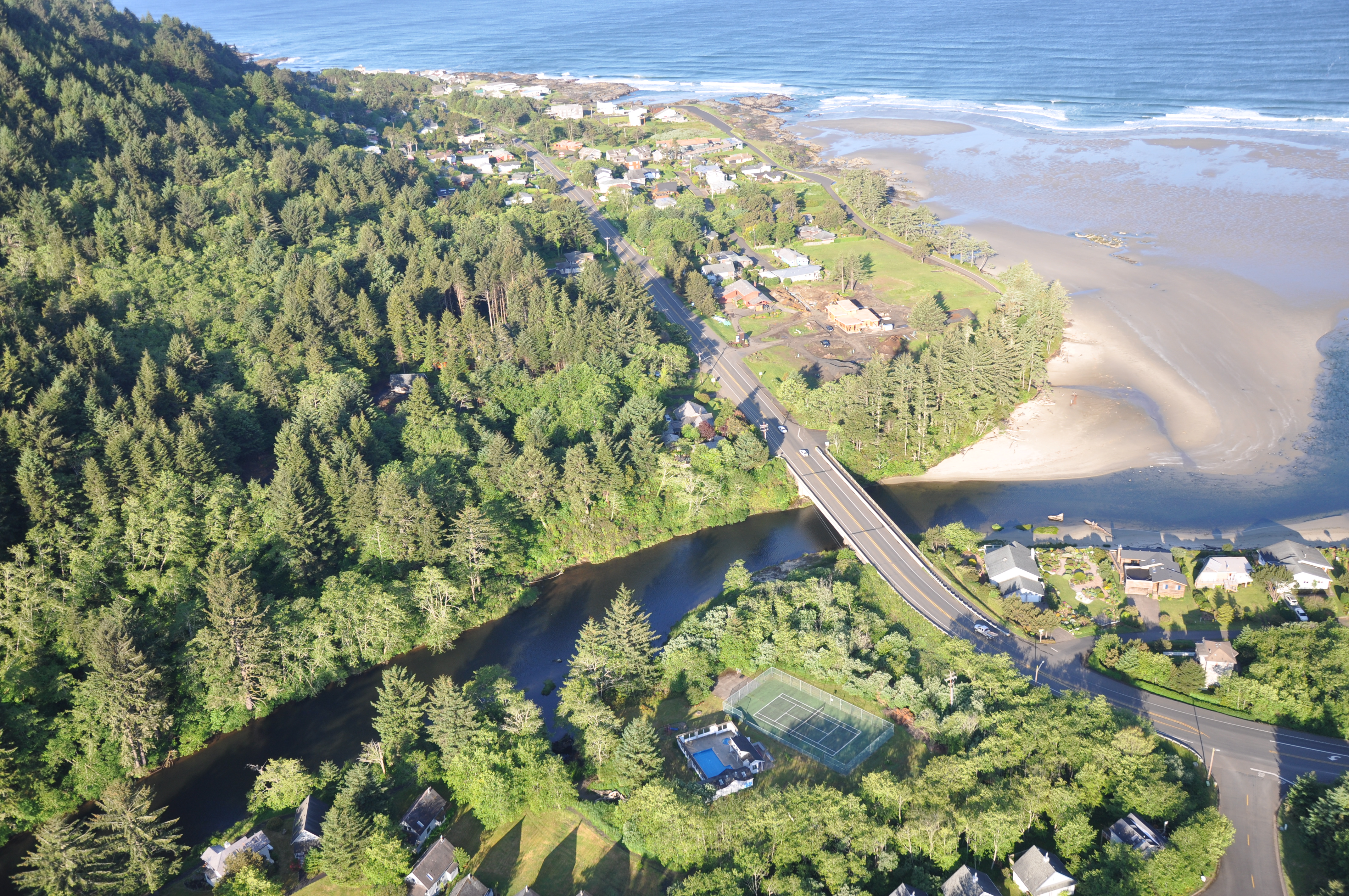 An aerial view of the Yachats River winding through dense pine trees, under a paved bridge, and into the ocean