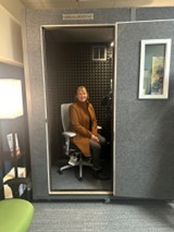 A picture containing Tina Ontiveros seated in a recording studio booth wearing a tan coat and smiling.