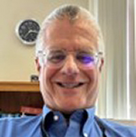Photo of Ian, a man with short grey hair and smiling at the camera. He is wearing a blue button-up collar shirt and thin rimmed glasses. 