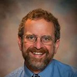 Photo of Jim, a man with short and curly red hair and beard with silver blended in, with glasses smiling at the camera
