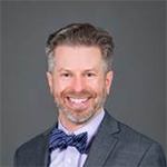 Photo of Jonathan, a man with short, wavy, light grey hair he is smiling at the camera. He is wearing a grey suit over a light lavender, button-up, collar shirt with a blue striped bowtie. 