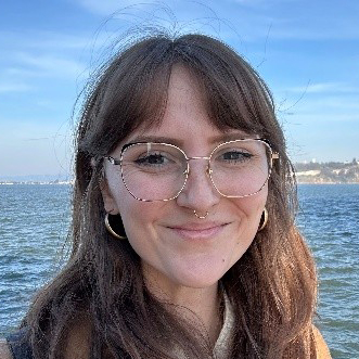 Photo of Kaitlin, a woman with shoulder length dark blonde hair. She is wearing a brown coat, gold rim glasses.