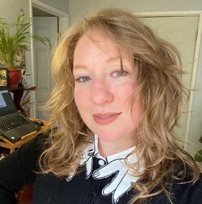 Katie, a woman with shoulder length wavy blonde hair, is smiling at the camera. She is wearing a black button up shirt with a white collar. The background is her home office setting