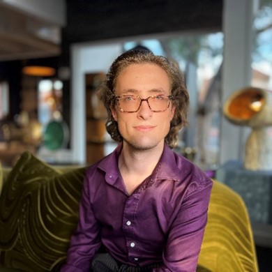 Andrew, a man with short light brown hair, is looking at the camera and is wearing a purple button up shirt and sitting on an olive-green couch