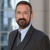 John, a man with short brown hair and beard is smiling at the camera and is wearing a light blue dress shirt, dark colored tie, and a black suit coat. The background is a gray wall over his left shoulder and a window with a blurred vision of a structure over his right shoulder