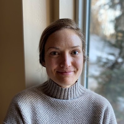 Katie, a woman with shoulder length brown hair that is pinned up is wearing a grey sweater with a high neck collar and smiling at the camera. The background is a cream color wall from center to left and a window with trees from center to right.