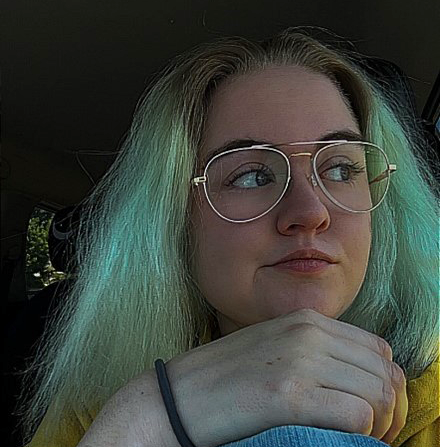 Olivia, a woman with medium length light brown and green hair and glasses, is sitting with a knee up while looking towards her left. She is wearing blue jeans and a yellow shirt
