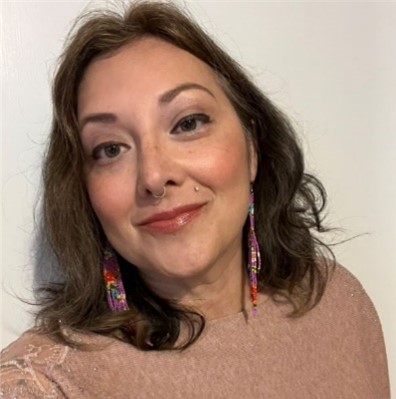 Rebecca, a woman with shoulder length brown hair, is wearing a champagne colored sweater and dangly earrings and is looking at the camera