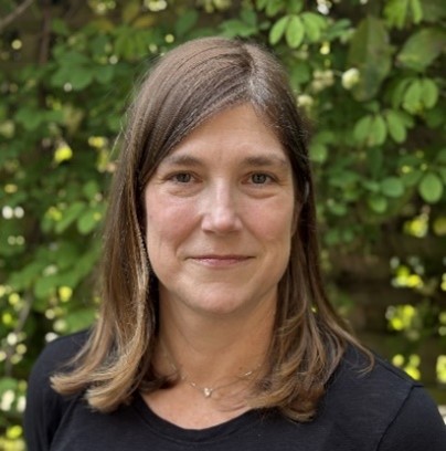 Suzanne, a woman with shoulder length brown hair is smiling at the camera and is wearing a black shirt. The background is green leafy trees.