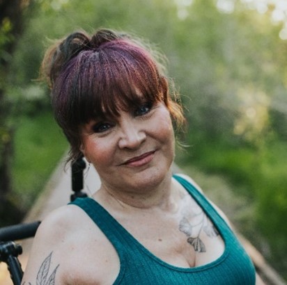 Corinne, a woman with long brown hair with red highlights pinned up, is smiling at the camera. She is wearing a green tank-top with a tattoo on her upper right arm and left chest. The background is a nature scene.