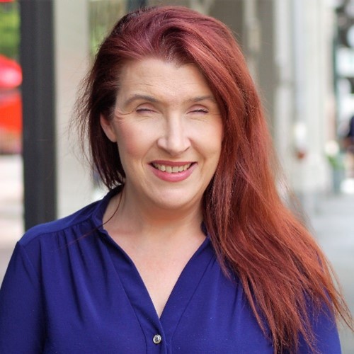 Emily, a woman with long red hair, is wearing a royal blue button up shirt and smiling at the camera. The background is a city scape.