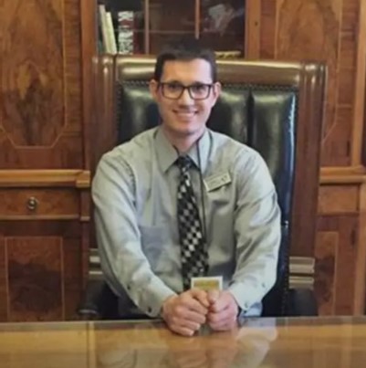 Eric, a man with short black hair and glasses is smiling at the camera. He is wearing a grey dress shirt, grey tie, and sitting in a black office chair. The background is office cabinetry.
