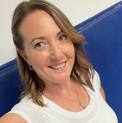 Jennifer, a woman with shoulder length light brown hair and she is smiling at the camera. She is wearing a white shirt with a silver necklace. The background is a white wall with a blue mat.