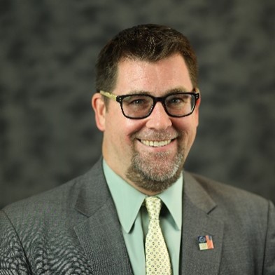 John, a man with short brown hair, light grey mustache and goatee, glasses is smiling at the camera. He is wearing a mint green dress shirt, green and white necktie, and grey suit coat with an American Flag pin. The background is a grey photo screen.