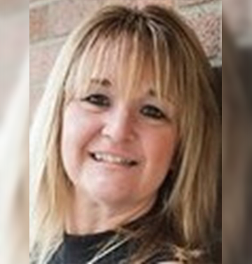 Kim, a woman with shoulder length blonde hair smiling at the camera. The background is a brick wall.