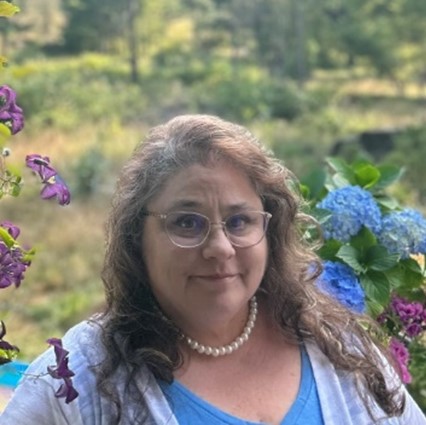 Melanie, a woman with long light brown hair with glasses is looking at the camera. She is wearing a blue V-neck top with a white shawl and white necklace. The background is a nature scene.