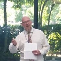 Ted, a man with a bald head, glasses, and short beard, is holding a paper in his left hand and giving a speech at the camera. Ted is wearing a white button up shirt with a brown tie. The background is glass windows with trees and shrubbery outside.