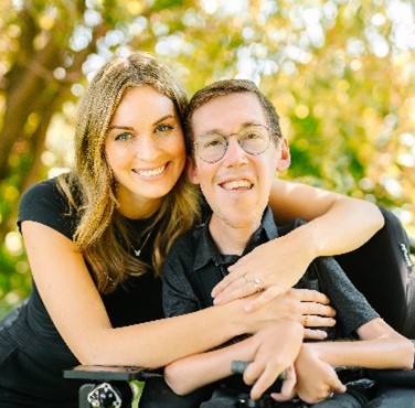 Hannah is a woman with long blonde hair smiling at the camera with her arms around Shane. Shane is a man with short brown hair, glasses, sitting in his power wheelchair, and smiling at the camera. The background is trees and the glow of the sun.