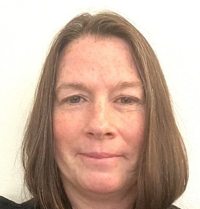 Brenda, a woman with shoulder length light brown hair is wearing a black shirt and looking at the camera. The background is a white wall.