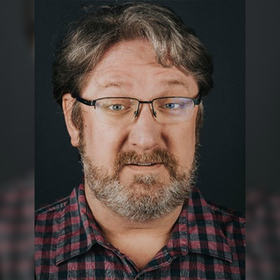 Todd, a man with short brown hair, brown and silver beard, and black framed glasses is looking at the camera. He is wearing a black and red checkerboard button up shirt. The background is a black wall.