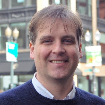 Tom, a man with short brown hair, is smiling at the camera. He is wearing a blue pull over on top of a light blue collared shirt. The background is a city scene.