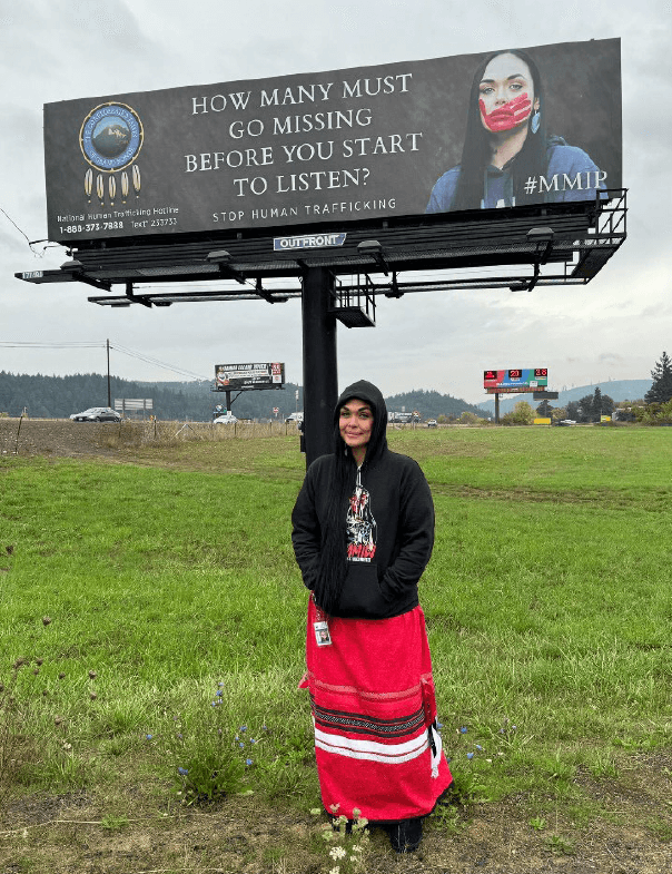 Amanda Freeman wears a black hoodie and red ribbon skirt, standing in front of a Missing and Murdered Indigenous People billboard