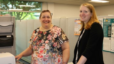 Two employees standing inside an office and smiling at the camera