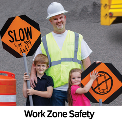 A work zone flagger holds 2 children who hold signs that say: Slow for my dad and no cellphones.