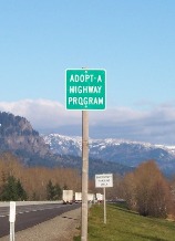 A green Adopt-A-Highway sign on the right side of a road