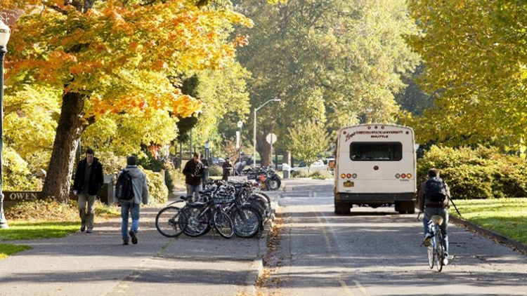 A photo representing transportation alternatives, including rear view of a shuttle bus, biker, and pedestrians on a fall day at Oregon State University.