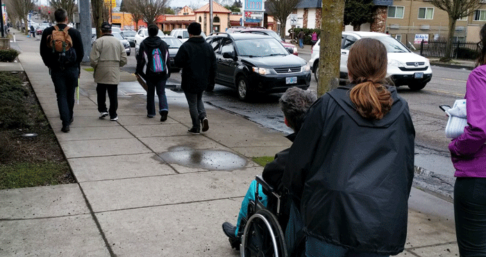 A group of pedestrians, with one man in a wheelchair, take a tour of 82nd Avenue.