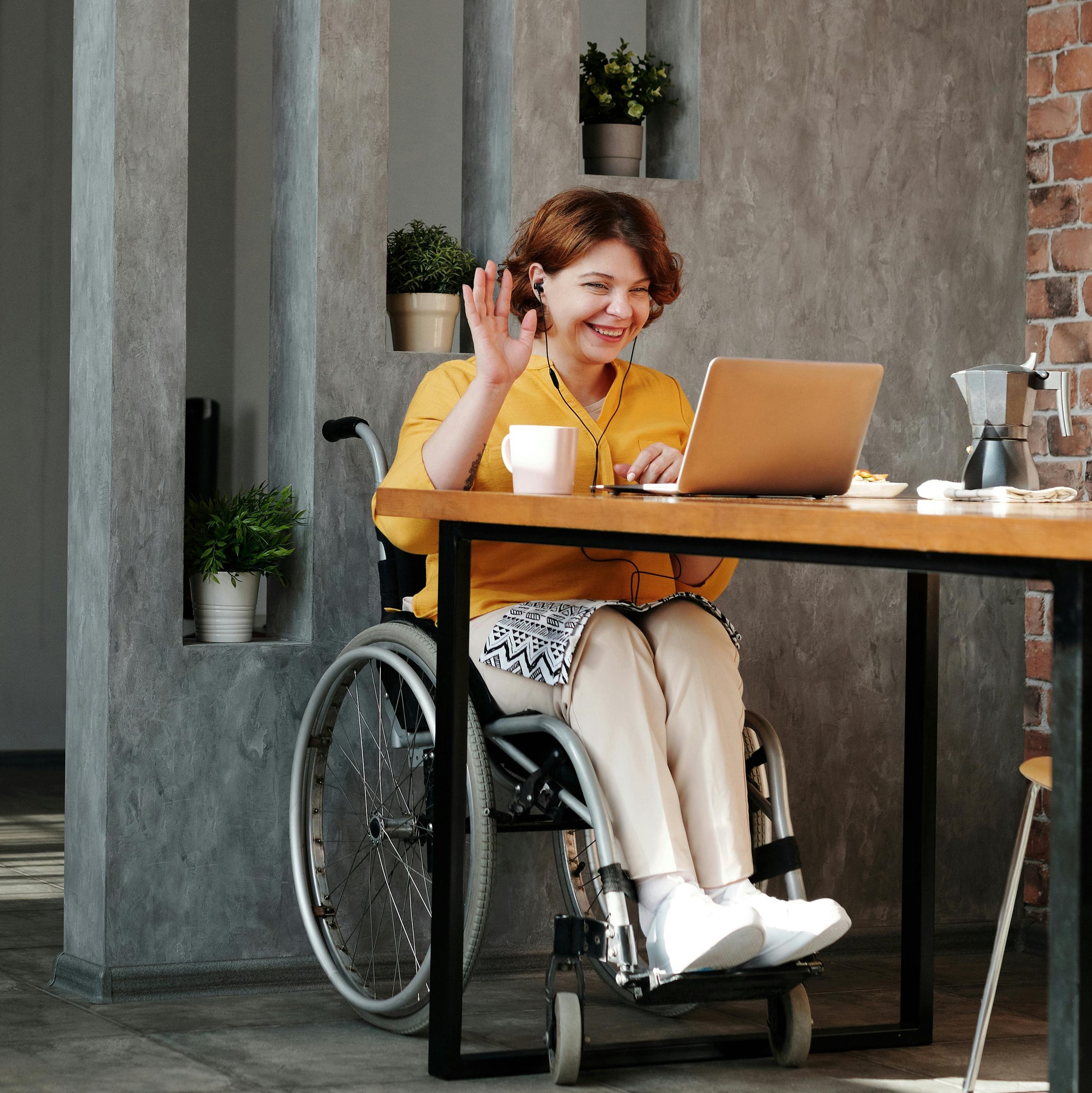 Image of woman in a wheelchair at table on conference call, waving at the camera with cup of coffee beside her.