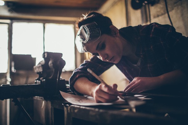 A worker in a machine shop using a pencil on top of their equipment. They have protective googles on their head.