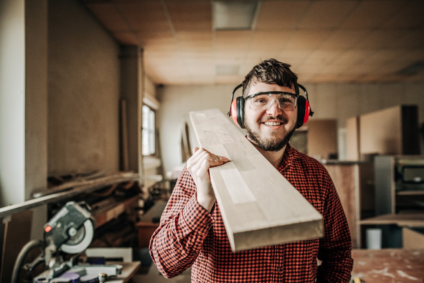 A young worker wearing safety glasses and ear protection holds a wooden board.