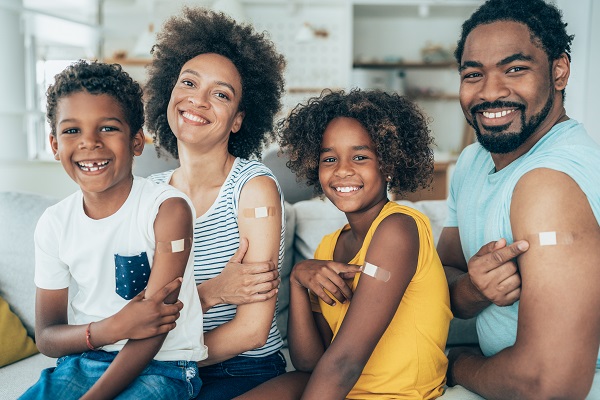 Smiling family of four sitting next to each other pointing at bandaids on their right arm.